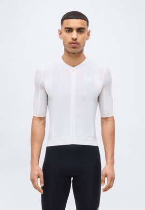 Jonge man staat recht vooruit, gekleed in een nauwsluitend wit fietsshirt met korte mouwen en zwarte fietsshorts.