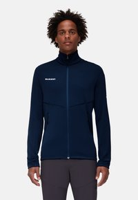 Marinblå fleecetröja med dragkedja och hög krage, kontrasterande övre panel och två fickor med dragkedja på sidan. Logotyp syns på bröstet.