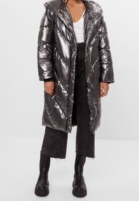 Skinande, metallisk grå puffjacka med huva, med ett chevronmönster och långa ärmar, bärs över svarta vida byxor och kraftiga svarta stövlar.