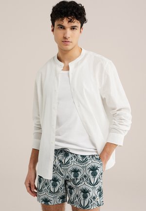 Jeune homme portant une chemise blanche à boutons par-dessus un T-shirt blanc et un short à motifs avec un cordon de serrage, debout avec une main dans la poche.