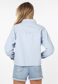 Světle modrá džínová bunda s dlouhými rukávy, límečkem a třepeným lemem, ve spojení s džínovými šortkami. Jednoduchý, neformální design, bez viditelného loga.