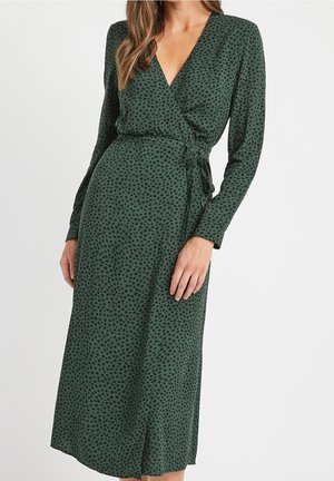 Donna che indossa un abito verde a portafoglio con maniche lunghe, motivo a puntini neri irregolari e cintura annodata sul lato, in piedi davanti a uno sfondo chiaro uniforme.