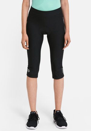 Person trägt schwarze, gepolsterte Radhosen, die unterhalb der Knie enden, und schwarze Sportschuhe, steht vor einem schlichten Hintergrund.