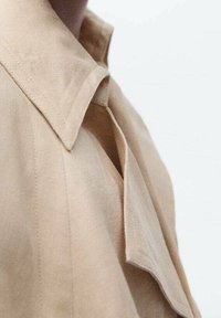 Chemise en lin beige avec un col évasé, présentant des coutures visibles et une coupe ample. Le tissu semble doux avec un léger éclat.