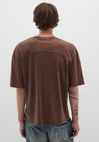 Maglietta marrone a maniche corte con vestibilità rilassata, dettagli a cucitura piatta sulle spalle e tessuto morbido. Indossata con jeans in denim.