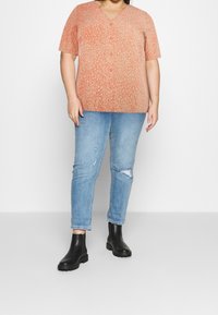 T-shirt à manches courtes en corail avec un motif beige ; associé à un jean déchiré bleu clair et des bottines noires.
