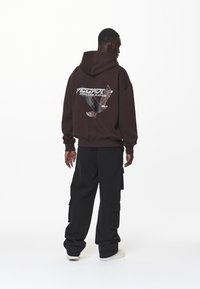 Felpa marrone con una grafica di un'aquila e il testo "SILENZIO SENZA FINE" sul retro, abbinata a pantaloni cargo neri e scarpe bianche.