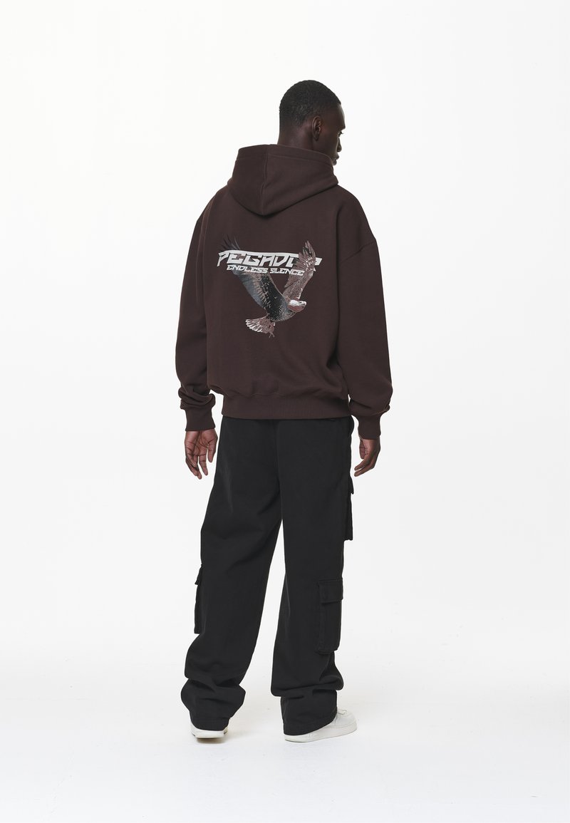 Felpa marrone con una grafica di un'aquila e il testo "SILENZIO SENZA FINE" sul retro, abbinata a pantaloni cargo neri e scarpe bianche.