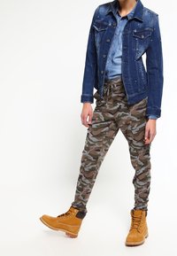 Jeansjacka med nitar, ljusblå skjorta, kamouflage-joggingbyxor och bruna stövlar. Fokusera på praktisk design och jordnära färgpalett.