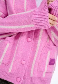 Růžový pletený cardigan s bílými pruhy, zdobený knoflíky ve tvaru srdce, ribbed manžetami a předními kapsami. Štítek viditelný na kapse.