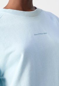 Sudadera azul claro con una textura suave, cuello acanalado y un discreto bordado de logo en la parte frontal. Diseño casual y ajuste relajado.