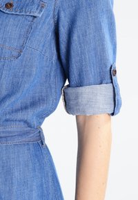 Rullattu sinisen farkkukleidin hihansuu, jossa on napitusyksityiskohta, henkilön käsivarren päällä tasaisella taustalla.