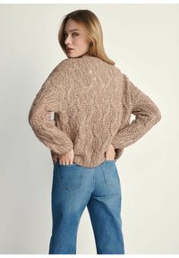 Gruba dzianinowa sweter w jasnobrązowym kolorze z teksturowanym wzorem w warkocze. Luźny fason, duże rękawy i ściągacz na dole. W zestawie z niebieskimi dżinsami.