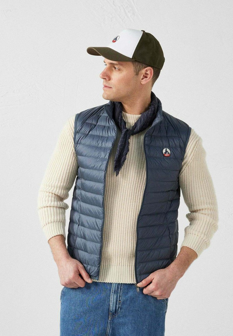Gilet rembourré gris sur un pull crème, associé à une écharpe marine. Le mannequin porte une casquette blanche et verte avec un logo. Un jean clair complète la tenue.