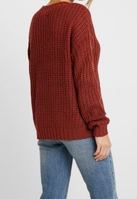 Sweter dziergany w bogatym, rdzawym kolorze z fakturowanym wzorem, ściągaczami na rękawach i u dołu; zestawiony z jasnoniebieskimi jeansami.