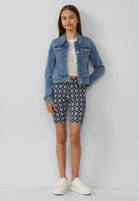 Spijkerjas met knoopsluiting, lichtblauwe kleur. Witte cropped t-shirt en patroon van marineblauwe shorts met ruitvormen. Witte sneakers.