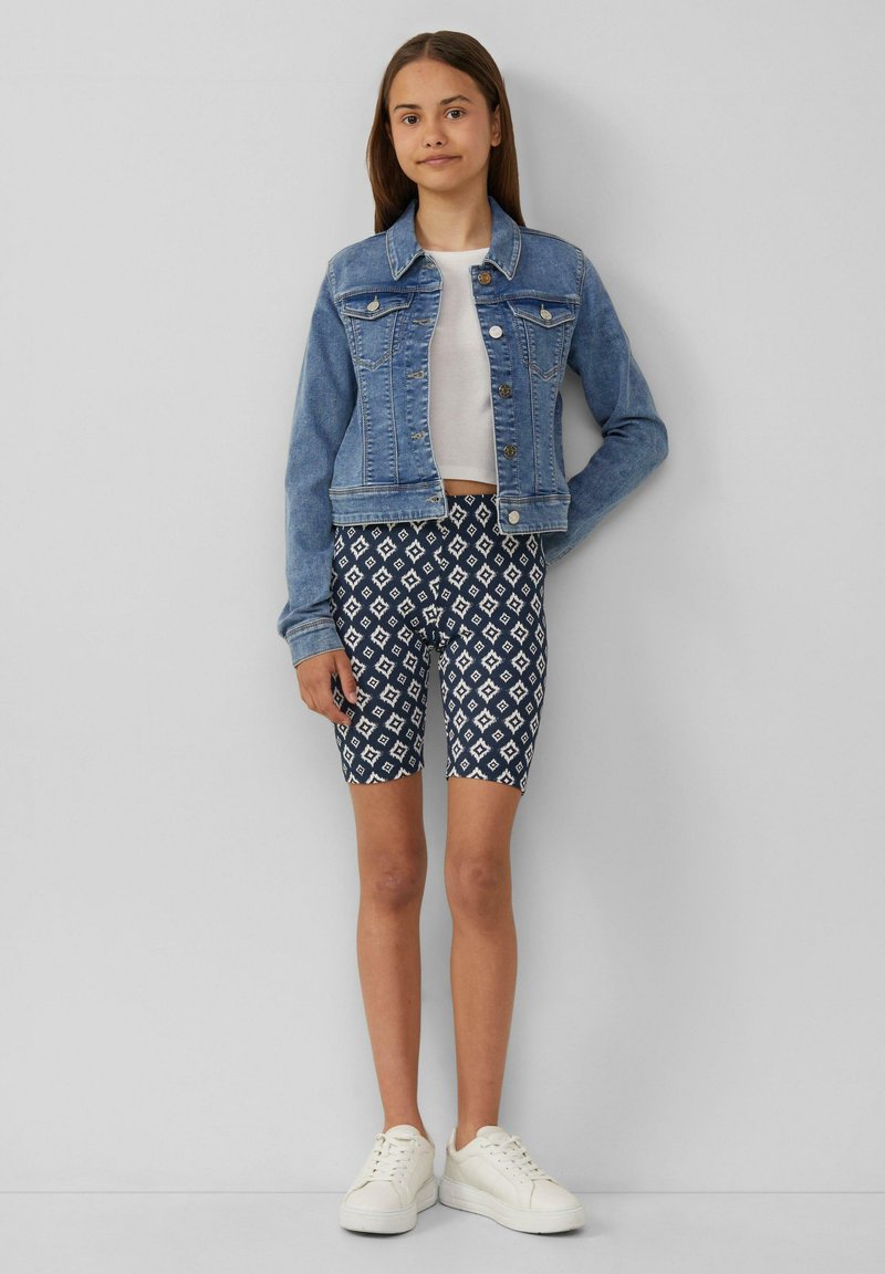 Spijkerjas met knoopsluiting, lichtblauwe kleur. Witte cropped t-shirt en patroon van marineblauwe shorts met ruitvormen. Witte sneakers.