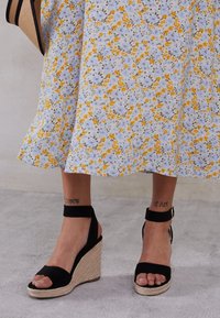 Black wedge sandals with a fabric upper, ankle strap, and woven jute platform, paired with a floral print dress in yellow and blue tones.