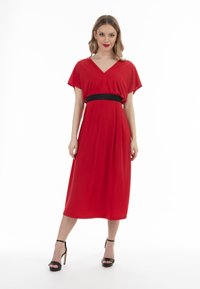 Rode midi-jurk met korte, losse mouwen, een V-hals en een zwarte textuurriem op de taille. Draagt zwarte sandaaltjes met straps.