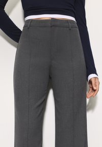 Grijze, op maat gemaakte broek met een gladde textuur, rechte pijpen en een voorste plooi. De tailleband zit net boven het heupniveau.