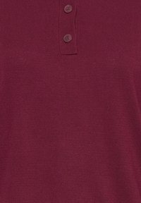 Gros plan sur un tissu tricot bordeaux avec un col côtelé et deux boutons ronds assortis sur la patte de boutonnage.