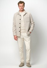 Giacca camicia beige a spina di pesce con bottoni neri, indossata sopra un maglione chiaro e pantaloni crema. Sneakers grigie completano l'outfit.