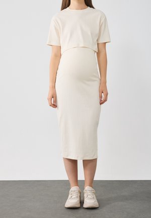 Zwangere vrouw die een roomkleurige ribgebreide jurk met korte mouwen en beige sneakers draagt, staand op een grijze vloer tegen een witte achtergrond.