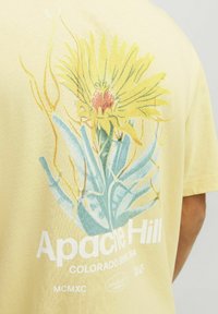 T-shirt di cotone giallo con un design grafico floreale in verde e giallo, con la scritta "Apache Hill" in lettere bianche in grassetto.