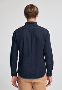 Mörk denimskjorta med långa ärmar, knappad krage och kontrasterande sömmar. Slät textur med böjd nederkant.