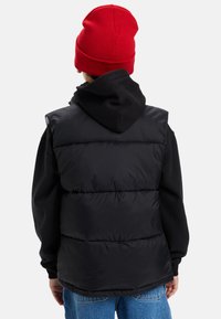 Person med en svart pufferväst över en svart hoodie, blå jeans och en röd stickad mössa, vänd bort från kameran mot en vit bakgrund.