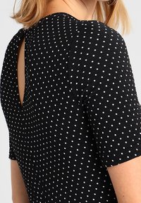 Svart topp med prickar och korta ärmar, med en texturerad tyg och en V-formad ryggutskärning. Vita prickar är jämnt fördelade över hela toppen.