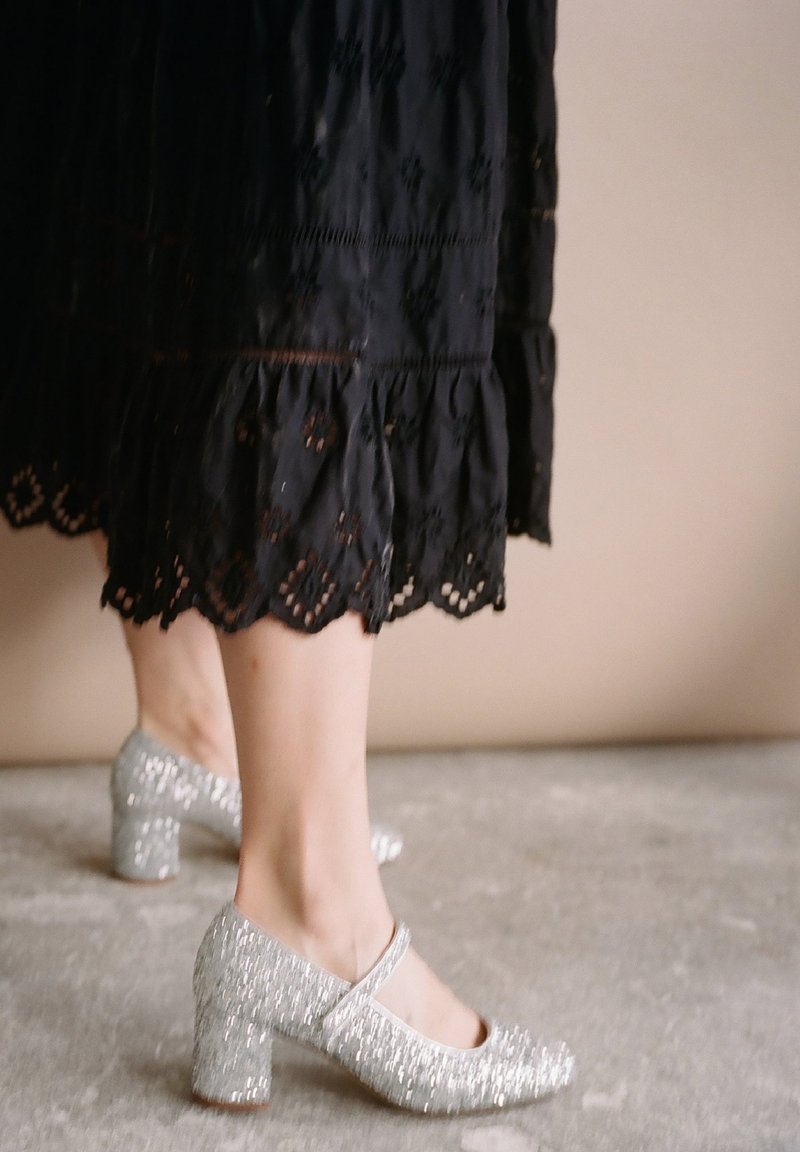 Zapatos de tacón alto plateados con brillo y correas en el tobillo, con un tacón grueso y punta redonda, combinados con un vestido negro bordado.