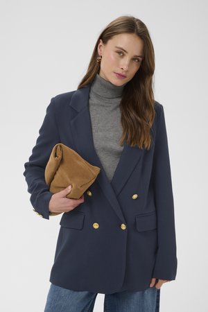 Hosszú barna hajú nő szürke garbót és tengerészkék blézert, arany gombokkal viselve, összehajtott bézs színű velúr borítéktáskát tartva.