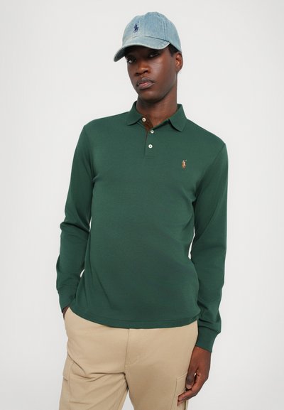 Camisa polo de manga larga verde con un cuello marrón y tres botones; presenta un logo bordado en el pecho, acompañada de una gorra de mezclilla.