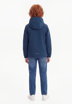 Enfant aux cheveux roux bouclés portant une veste à capuche bleu marine, un jean bleu et des chaussures de sport blanches, debout de dos sur un fond blanc uni.