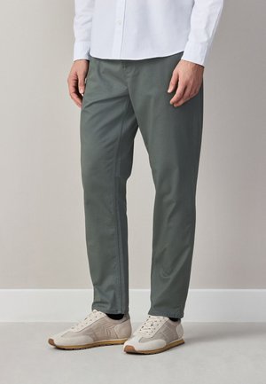 Egy férfi szürke slim fit nadrágot, fehér inget és bézs tornacipőt visel, beltéren áll egy világos fal és padló előtt.