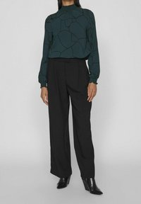 Blusa a maniche lunghe verde scuro con un motivo astratto nero, abbinata a pantaloni neri a vita alta e stivaletti neri con tacco a punta.
