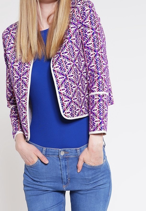 Femme portant une veste courte à motifs violets, un haut ajusté bleu et un jean bleu, les mains dans les poches avant, sur un fond uni.