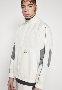 White, lightweight jacket with gray accents, zip closure, and elastic cuffs. Features "AIR" logo in black and gold on the chest.