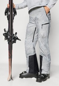 Pelēki slēpošanas bikses ar struktūru audumu un melniem akcentiem, ar vairākiem rāvējslēdzēju kabatām un logotipu. Slēpošanas aprīkojums turēts vienā rokā.