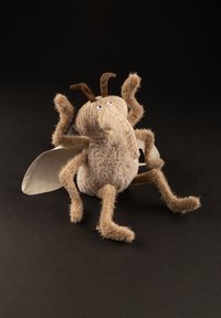Plush insect toy with a beige fuzzy body, two large eyes, textured legs, soft antennae, and beige wings, set against a dark background.