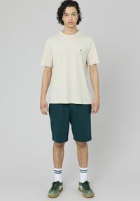 T-shirt en coton beige avec un petit motif imprimé, associé à un short vert foncé. Le modèle porte des chaussettes blanches avec des rayures vertes foncées et des baskets vertes.