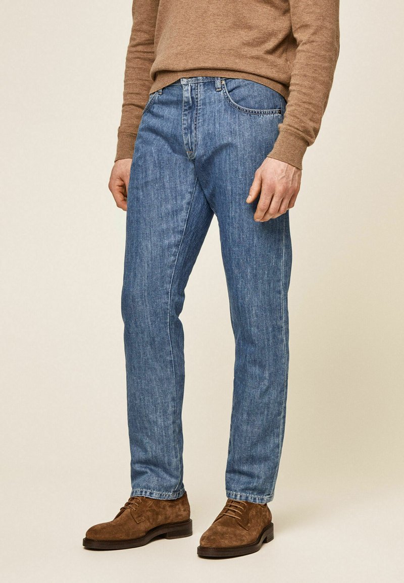 Jeans de denim azul con un corte recto, que presentan un diseño clásico de cinco bolsillos y un sutil desgaste. Combinados con zapatos de ante marrones y un suéter.