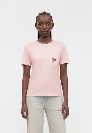 Vrouw met gevlochten haar die een lichtroze T-shirt draagt met een klein bloemenpatroon op de linkerborstzak en lichtgrijze gescheurde spijkerbroek.