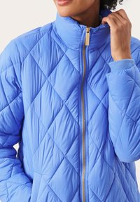 Jasnoniebieska pikowana kurtka z wysokim kołnierzem, złotym zamkiem i ściągaczami. Teksturowany wzór diamentów na całej tkaninie.