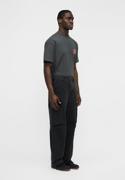 Γκρι βαμβακερό μπλουζάκι με κοντά μανίκια και μικρό κόκκινο σχέδιο στο στήθος, σε συνδυασμό με χαλαρής γραμμής μαύρο cargo παντελόνι με πλαϊνές τσέπες.