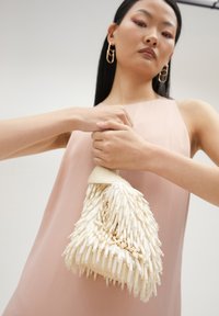 Biała torebka o teksturowanej powierzchni i kształcie stożka, z koralikową powierzchnią i plecionym paskiem. Model trzyma torbę na jasnym tle.