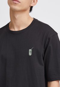Black cotton t-shirt with a round neckline, featuring a small, textured graphic of a drink can with a straw on the left chest area.