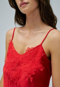 Camisola de satén roja con delicados detalles de encaje en el escote. Cuenta con tirantes finos ajustables y una textura suave.