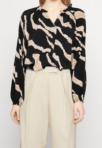 Blusa nera a maniche lunghe con motivo astratto beige, dettaglio nella scollatura arricciata, vestibilità ampia e polsini elastici, abbinata a pantaloni beige a vita alta.